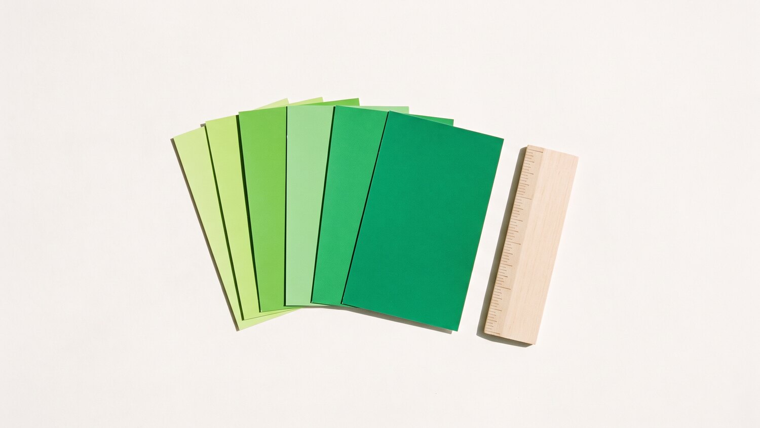 Six green paint swatch cards fanned across a pale surface beside a small wooden ruler