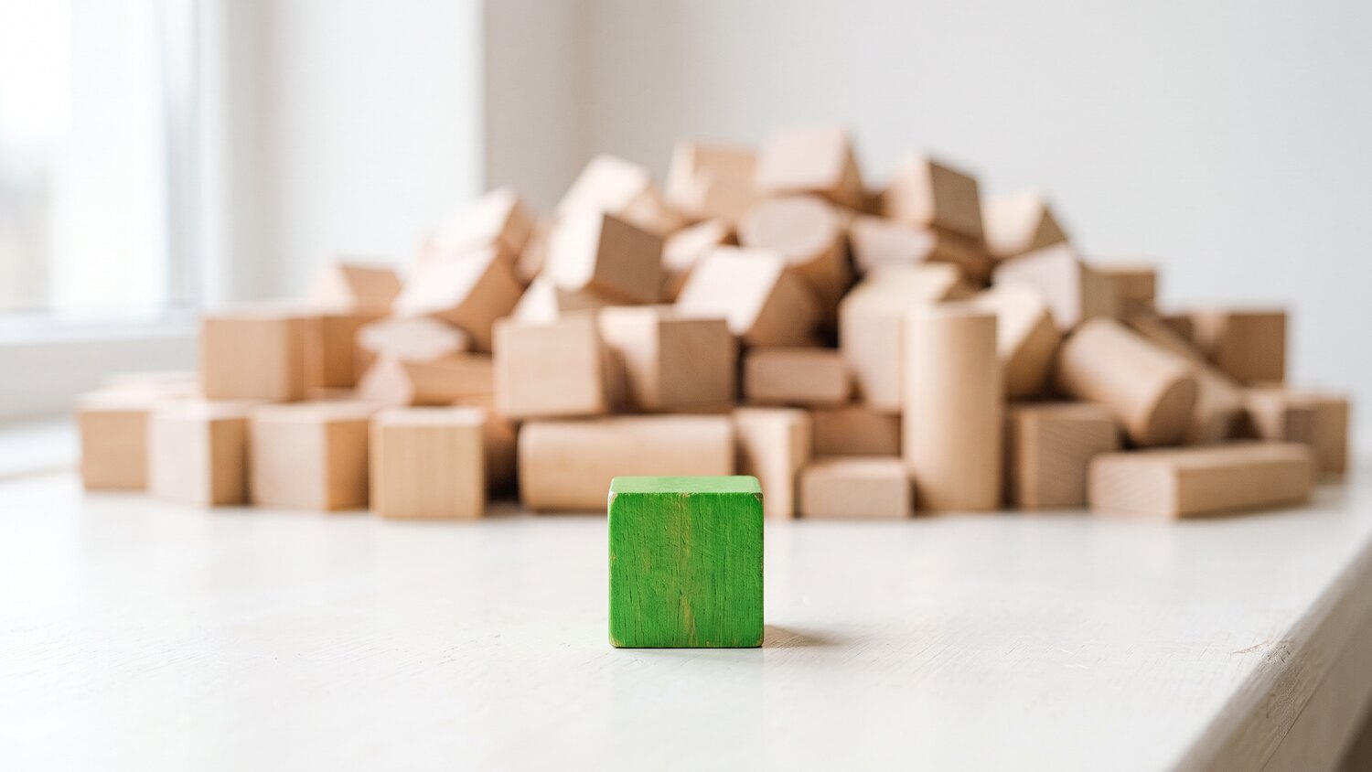 A single small green wooden block standing alone in front of a blurred heap of pale wooden blocks