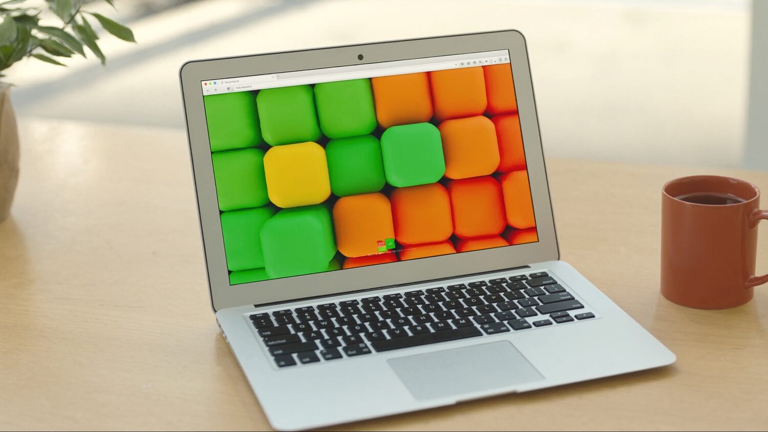 A laptop on a sunlit desk showing a colourful grid of product tiles, beside a mug and a plant