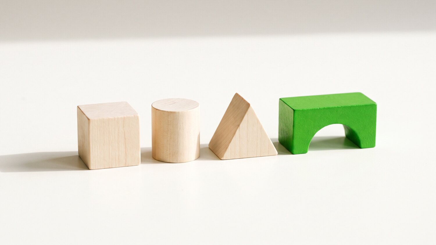 Four pale wooden blocks of different shapes in a row, a cube, cylinder and prism, with a green arch