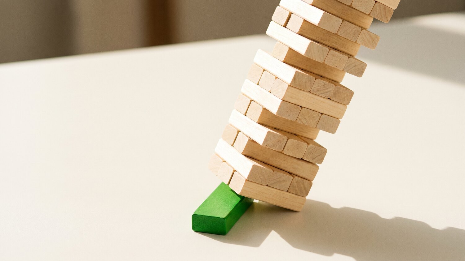 A tall leaning stack of pale wooden blocks with a single green block wedged near the base taking the weight